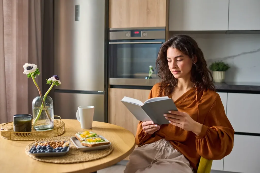 young relaxed woman in casualwear reading book while sitting by kitchen table